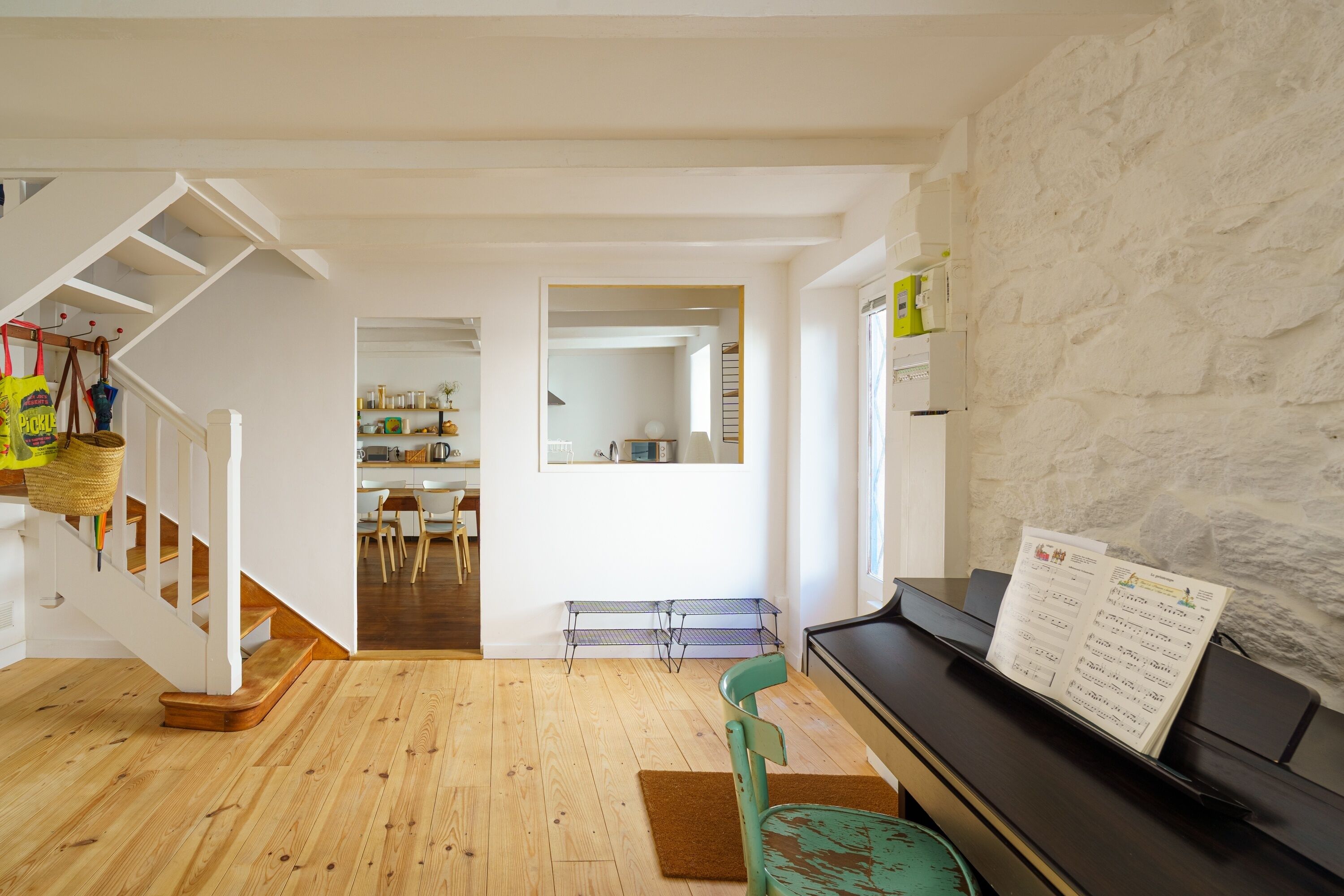 A spacious living room with a piano, wooden flooring, a staircase, and a kitchen in the background.