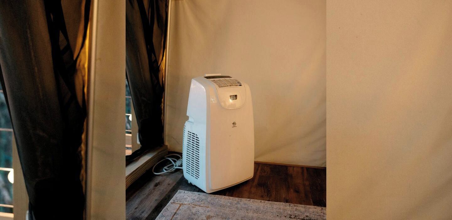 A white portable heater in a corner of a room with a wooden floor and a curtain partially drawn.