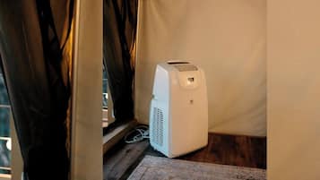 A white portable heater in a corner of a room with a wooden floor and a curtain partially drawn.