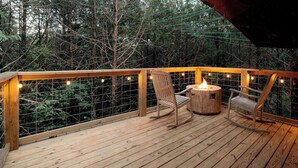 A wooden deck with a fire pit and two rocking chairs, surrounded by a wire mesh railing and string lights.