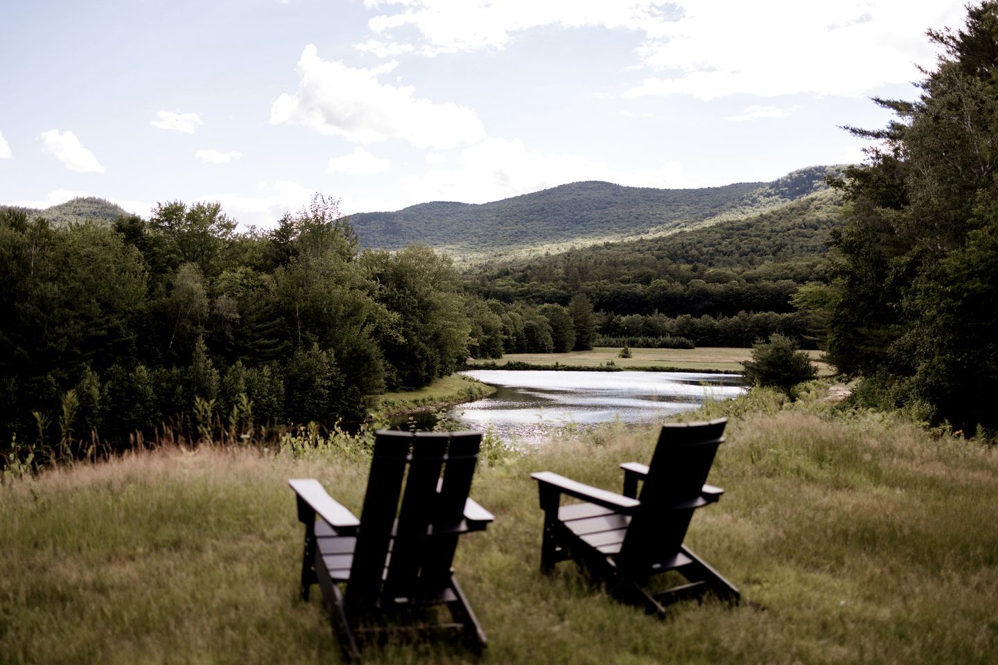 Two Adirondack chairs facing a river with a forested hillside in the background.