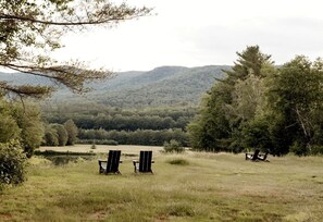 A natural landscape with two wooden chairs and a picnic table in a grassy field, surrounded by trees and hills.
