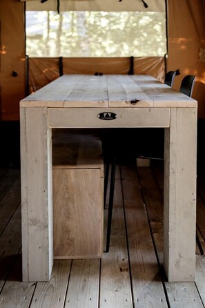 A wooden table with a drawer, set in a tent with a view of trees.