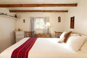 A bedroom with a large bed, a wooden headboard, a bedside table with a lamp, a small chair, and a window with curtains. - Studio-Style Log Cabin near Carson National Forest, New Mexico (San Cristobal)