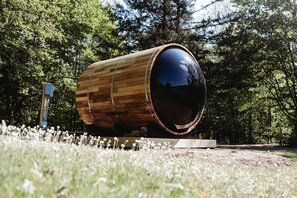 A large wooden barrel structure with a reflective surface, surrounded by a grassy area and trees.