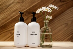 Deux flacons blancs étiquetés « Lotion pour les biens communs » et « Savon pour les mains » sont placés à côté d’un flacon en verre transparent contenant des fleurs.