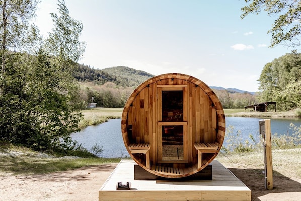 A wooden barrel-shaped structure with a bench inside, situated by a lake with a wooden deck in front.