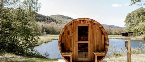 A wooden barrel-shaped structure with a bench inside, situated by a lake with a wooden deck in front.