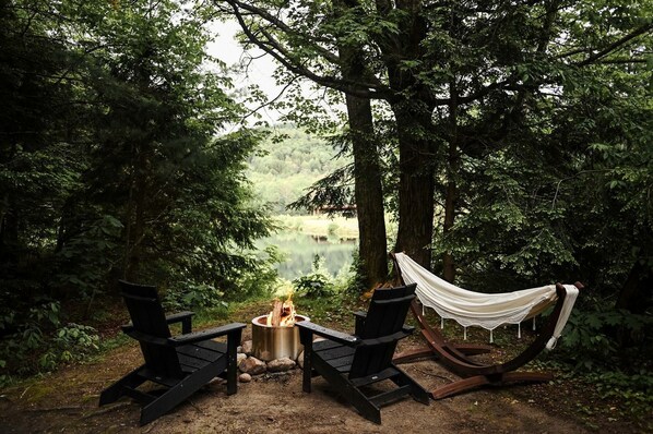 A hammock between trees, two chairs, a campfire, and a view of a lake.