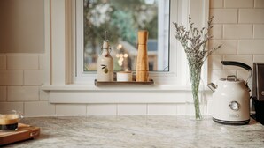 A kitchen counter with a window, a white kettle, a wooden pepper grinder, a glass vase with dried flowers, and a small candle holder.