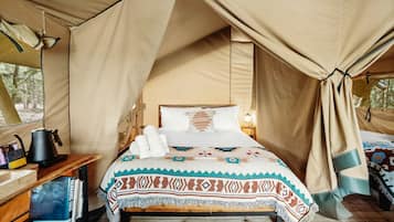 A tented room with a bed, pillows, a bedside table with a kettle, and a blanket.