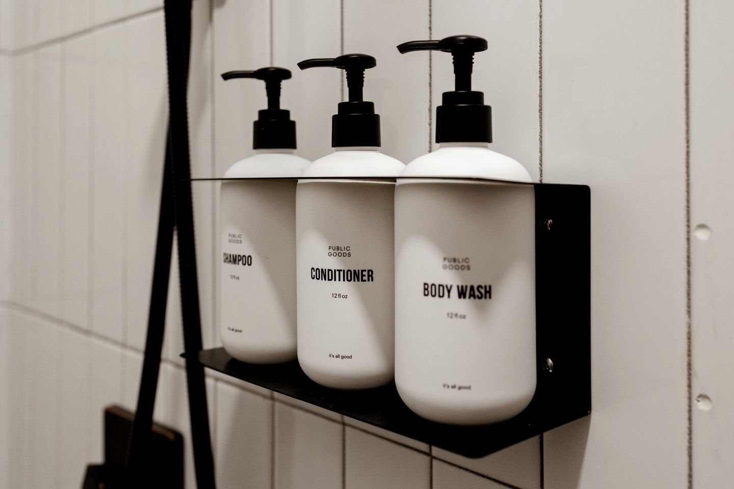 Ein schwarzes Regal mit drei weißen Flaschen, beschriftet mit 'Shampoo', 'Conditioner' und 'Body Wash', an einer weiß gefliesten Wand befestigt.
