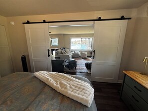Ein Schlafzimmer mit Bett, Schreibtisch und Blick in einen Wohnbereich mit Sofa und Poufs.