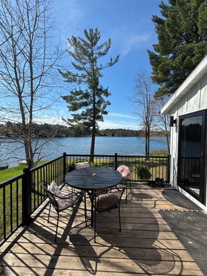 A wooden deck with a table and chairs, overlooking a lake with trees and a building in the background.