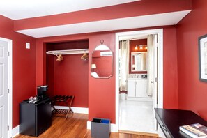 A hallway with a red wall, a mirror, a coat rack, a black speaker, a wooden bench, and a white door leading to a bathroom.