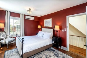 Un dormitorio con cama, dos lámparas de noche, una silla, ventana con cortinas, candelabro y puerta.
