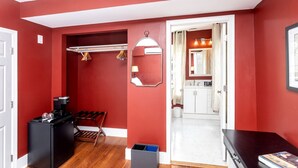 Un corridoio con una parete rossa, uno specchio, un appendiabiti, un altoparlante nero, una panca in legno e una porta bianca che conduce al bagno.