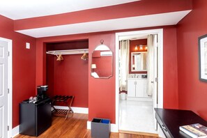 Un pasillo con pared roja, un espejo, un perchero, un altavoz negro, un banco de madera y una puerta blanca que da a un baño.