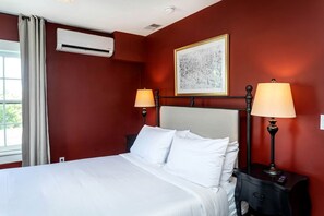 A hotel room with a bed, two bedside lamps, an air conditioning unit, a framed artwork on the wall, and a window with curtains.
