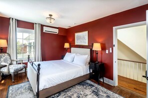 A bedroom with a bed, two bedside lamps, a chair, a window with curtains, a chandelier, and a door.