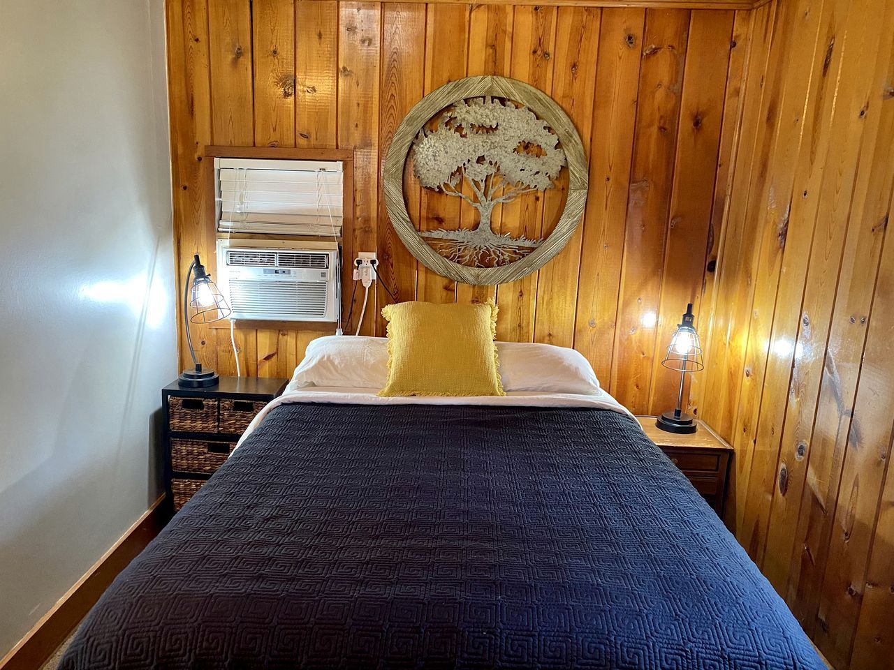 A bedroom with a wooden paneled wall, a bed with a patterned bedspread, a round wall decoration, a bedside lamp, and a window with blinds.