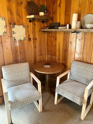 Une pièce d’angle comprenant deux chaises en bois, une petite table ronde et une étagère ornée d’objets décoratifs.