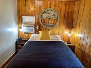 Ein Schlafzimmer mit einer holzverkleideten Wand, einem Bett mit gemustertem Überwurf, einer runden Wanddekoration, einer Nachttischlampe und einem Fenster mit Jalousien.
