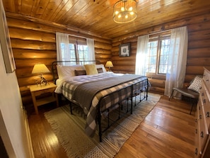 Un dormitorio de cabaña de madera con cama, mesitas de noche, cómoda y lámpara de araña.