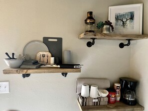 A kitchen with wooden shelves holding kitchenware, a coffee maker, and a framed picture.
