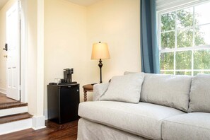 A room with a sofa, a coffee machine, a lamp, and a window with a view of trees.