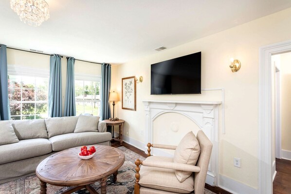 A living room with a sofa, armchair, round coffee table with fruit, a fireplace, and a TV mounted above it.