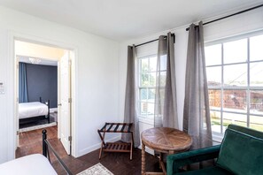 A room with a bed, a small table, a chair, a window with curtains, and a view of the outside.