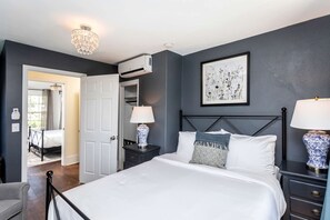 Una camera da letto con un letto, lampade da comodino, un comodino, una cassettiera, un lampadario, un condizionatore d'aria, una porta che dà su un'altra stanza e un'opera d'arte incorniciata appesa al muro.