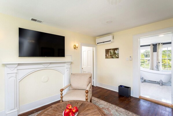 A living room with a fireplace, a television, a sofa, and a view of a garden through a window.