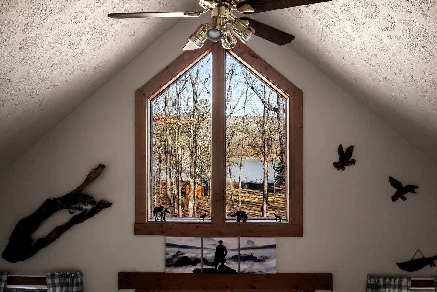 A bedroom with a sloped ceiling, a large window with a view of trees and a lake, a ceiling fan, and wall-mounted bird sculptures.