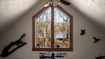 A bedroom with a sloped ceiling, a large window with a view of trees and a lake, a ceiling fan, and wall-mounted bird sculptures.
