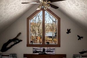 A bedroom with a sloped ceiling, a large window with a view of trees and a lake, a ceiling fan, and wall-mounted bird sculptures.