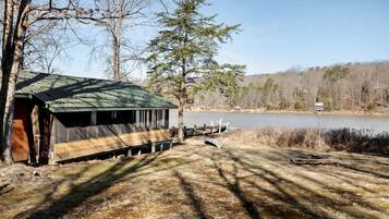 A rustic cabin with a green roof, situated by a lake with dock and boats, surrounded by trees and open fields.