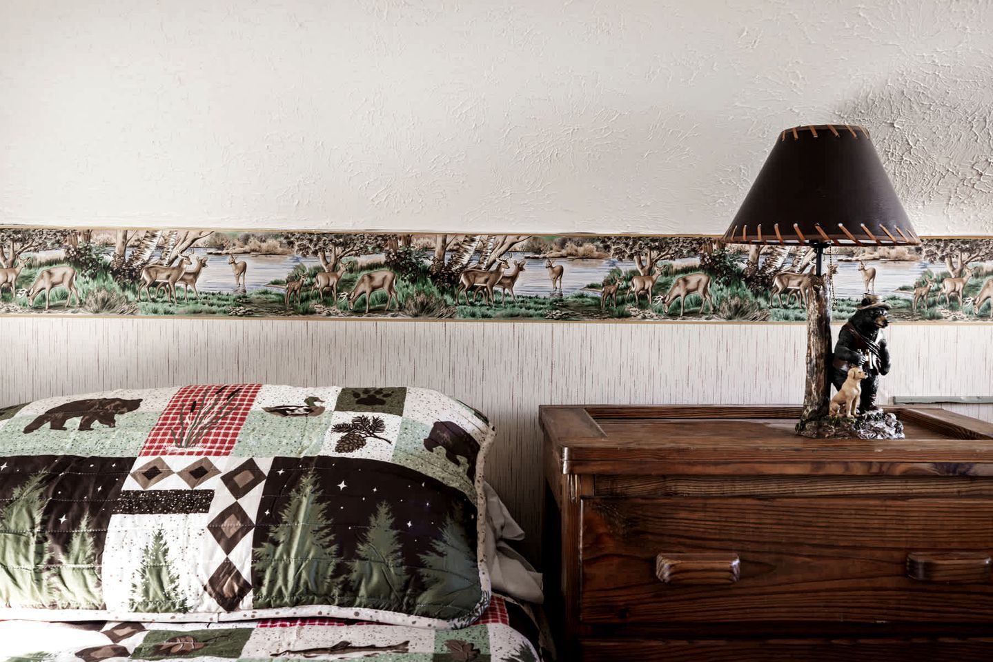 A bedroom with a bed, a wooden dresser, a lamp, and a wildlife-themed tapestry on the wall.