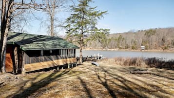 Un chalet rustique avec un toit végétalisé, situé au bord d’un lac doté d’un quai et de bateaux, entouré d’arbres et de champs ouverts.