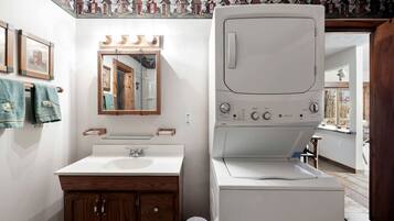 A compact bathroom with a white sink, wooden vanity, a mirror with lights, and a white washing machine.