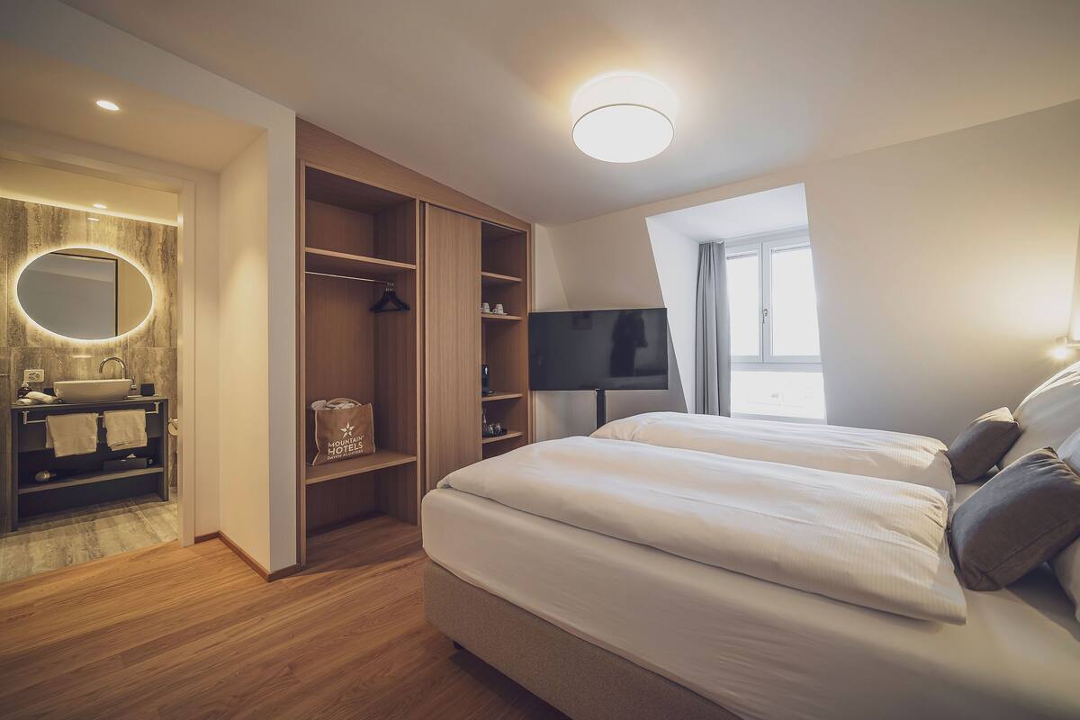 A hotel room with a bed, a TV, a wardrobe, a bathroom with a sink and mirror, and a window.