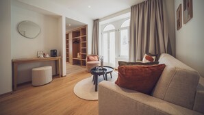 A modern living room with a sofa, coffee table, and a desk with a coffee machine. There is a closet with a hanging rack and a chair. The room has large windows with curtains. - Hotel Joseph's House (Davos)