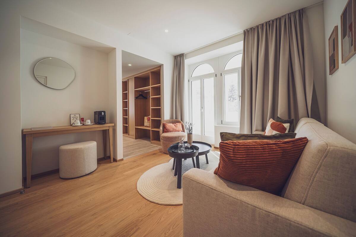 A modern living room with a sofa, coffee table, and a desk with a coffee machine. There is a closet with a hanging rack and a chair. The room has large windows with curtains.