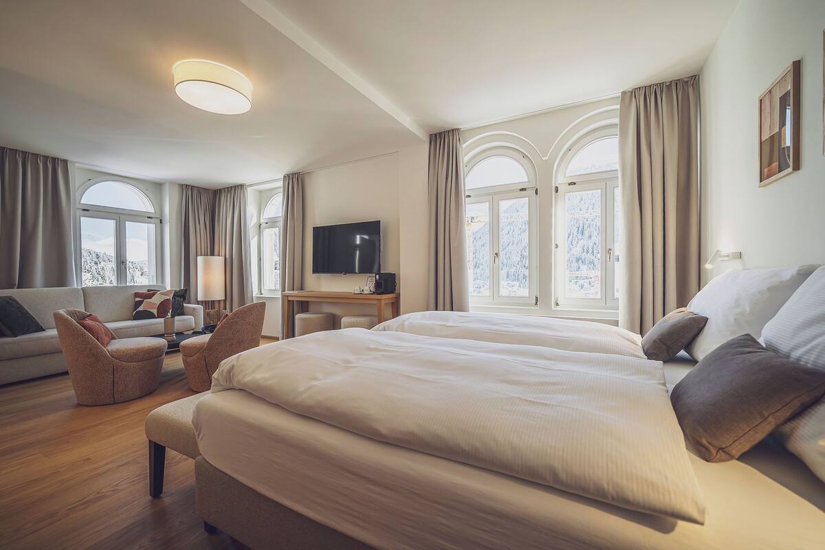 A hotel room with a large bed, a sofa, a desk with a TV, and a window with curtains.