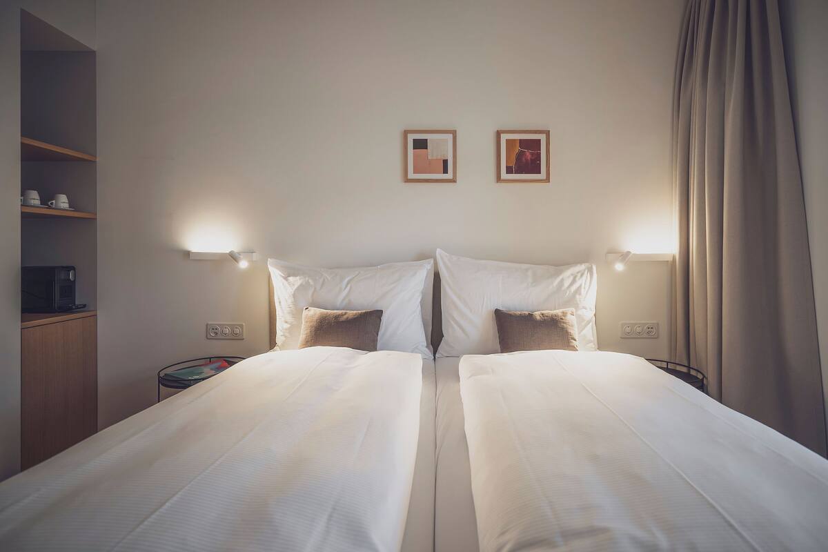 A double bed with white linens, two bedside tables, a shelf with cups, and two wall-mounted lights.