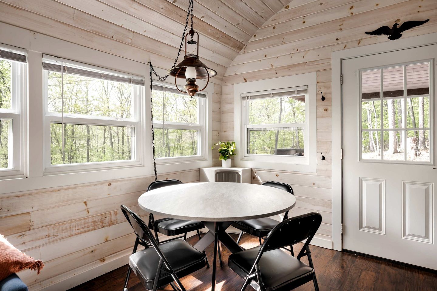 A room with a round table, four chairs, a hanging light fixture, and large windows with a view of trees.