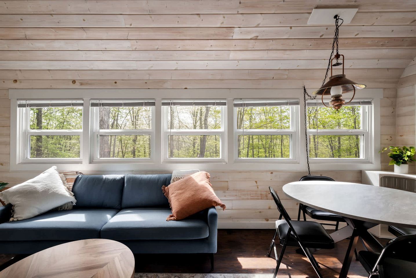 A cabin interior with a sofa, dining table, chairs, and large windows with a forest view.