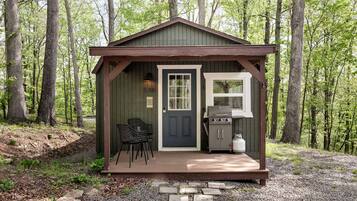 Une petite cabane avec une porte, une fenêtre, un barbecue et deux chaises à l’extérieur.