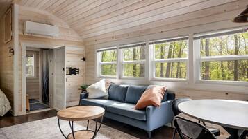 A cozy cabin interior with a wooden ceiling, a blue sofa, a round wooden table, and large windows offering a view of the forest.